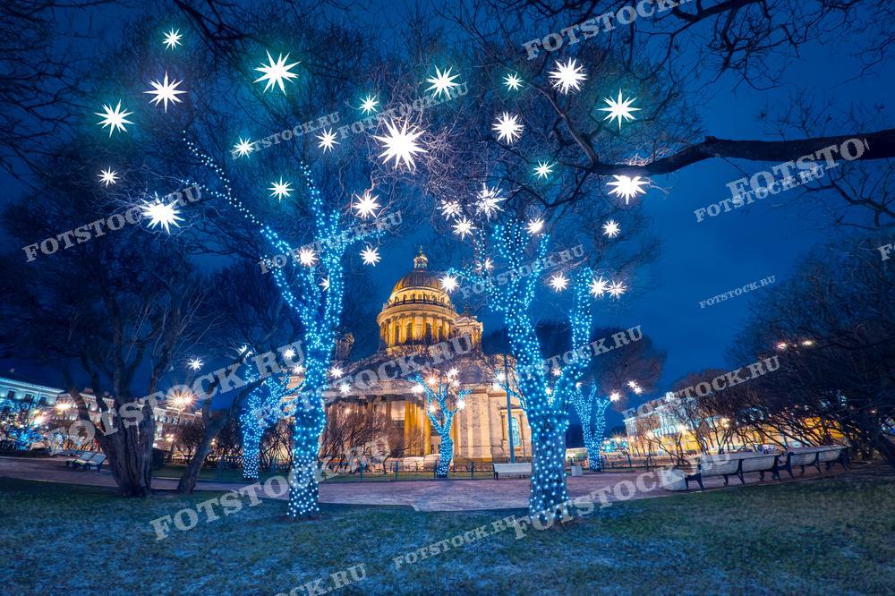 На фотографии изображен Исаакиевский собор в Санкт-Петербурге, окруженный новогодней иллюминацией. Деревья у собора украшены яркими гирляндами и световыми звездами, создавая волшебную атмосферу зимнего вечера. Величественный купол собора подсвечен, подчеркивая архитектурную красоту здания. Зимний Санкт-Петербург наполнен праздничным настроением.